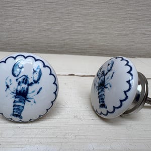 May include: Two white ceramic cabinet knobs with a blue lobster design. Each knob has a scalloped edge and a silver metal screw for installation. The lobster is depicted in a detailed, artistic style.