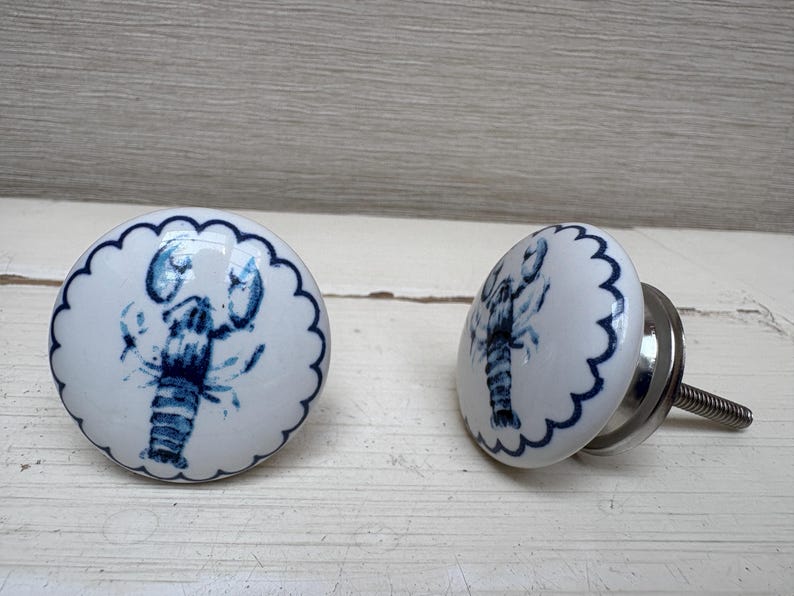 May include: Two white ceramic cabinet knobs with a blue lobster design. Each knob has a scalloped edge and a silver metal screw for installation. The lobster is depicted in a detailed, artistic style.