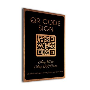 May include: A rectangular sign with a wood-grain border and a black centre. It features copper-coloured text reading QR CODE SIGN, Any Text, Any QR Code, and Double sided tape for easy (peel and stick) install, along with a central QR code graphic.