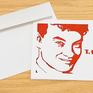 May include: A red and white greeting card with a red silhouette of Tom Hanks' face and the text "T. Hanks" in red.