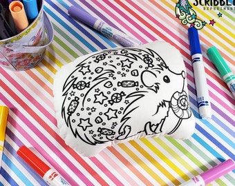 May include: A white plush hedgehog with a black outline, ready to be colored in. The hedgehog is surrounded by candy, stars, and swirls. The image includes a variety of colorful markers, including purple, blue, yellow, red, and pink.