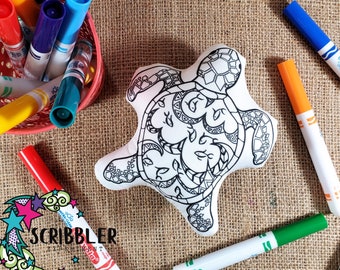 May include: A white turtle-shaped coloring pillow with black outline designs, surrounded by colorful markers. The markers are red, blue, orange, green, and black. The image includes the text "SCRIBBLER" with a star design.