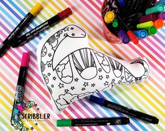 May include: A white fabric dinosaur cushion with a black and white line drawing of a dinosaur with stars, clouds, and a sun. The cushion is surrounded by colourful markers.