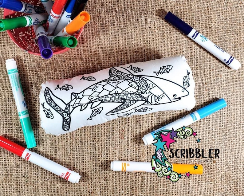 May include: A white pillow with a black and white line drawing of a shark and smaller fish. The pillow is surrounded by colorful markers.
