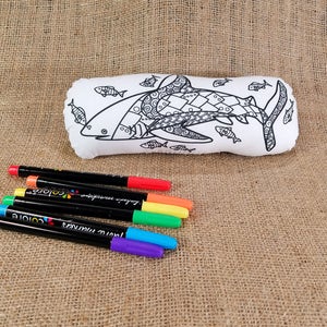 May include: A white fabric pillow shaped like a shark with a black and white patterned design. The pillow is surrounded by six colorful markers.