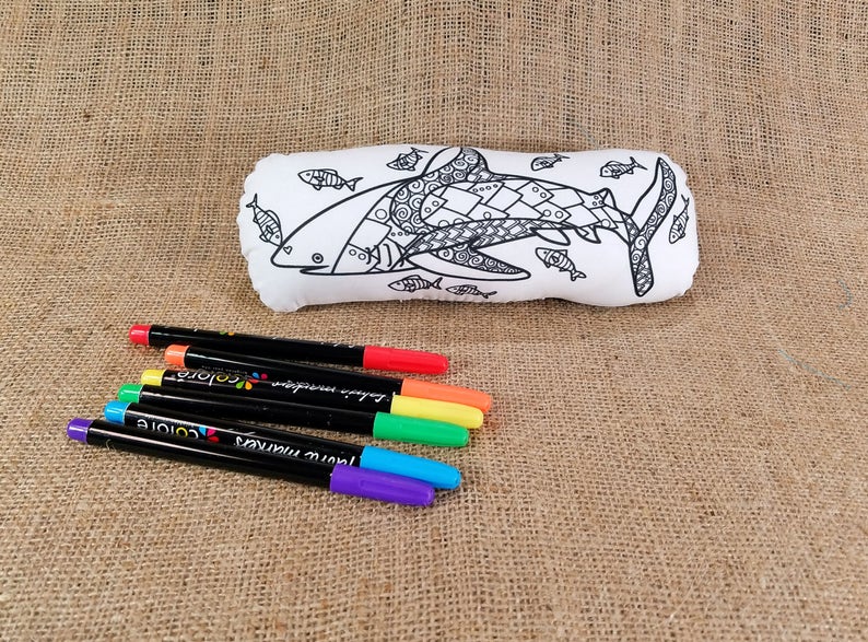 May include: A white fabric pillow shaped like a shark with a black and white patterned design. The pillow is surrounded by six colorful markers.