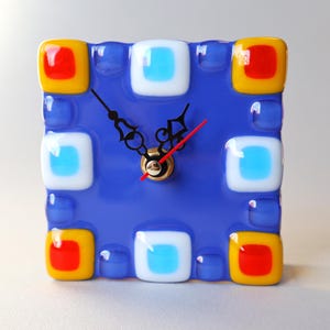 May include: A blue glass clock with red, yellow, and white squares around the face. The clock has black hands and a gold center.