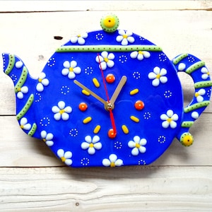 May include: Blue ceramic teapot clock with white daisy accents and yellow and orange details. The clock hands are black and red.