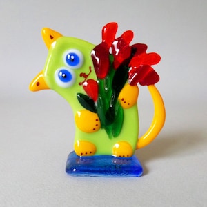 May include: A green glass cat figurine with yellow accents and large blue eyes, holding a bouquet of red flowers with green leaves. The cat is standing on a blue base.