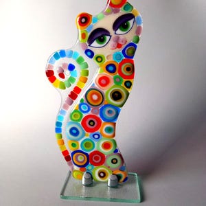 May include: A colorful glass cat figurine with a white face and large green eyes. The cat is covered in a pattern of multi-colored circles and is standing on a clear glass base.