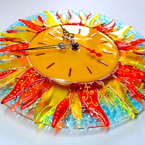 May include: A colorful glass clock with a sun design. The clock face is yellow with black hands and Roman numerals. The sun is made of red, orange, and yellow glass pieces.