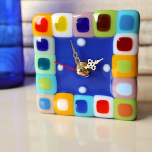 Puede incluir: Un reloj azul y blanco con un diseño de mosaico colorido. El reloj tiene una esfera blanca con puntos negros y manecillas doradas.