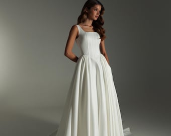 May include: A white satin wedding dress with a square neckline and a full skirt. The gown has a fitted bodice and a flowing skirt. The model is in a studio setting. The dress has a train.