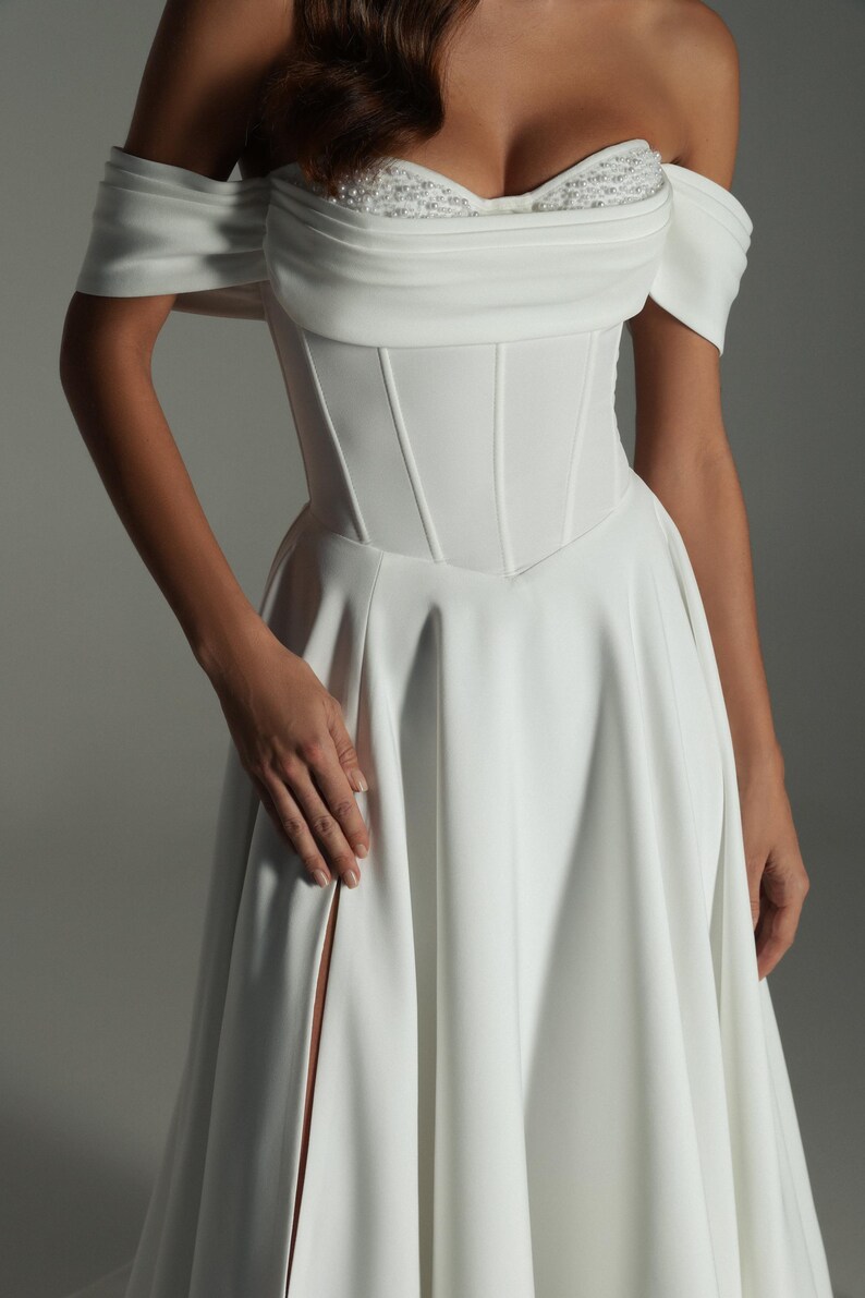 K&ouml;nnte beinhalten: Wei&szlig;es schulterfreies Brautkleid mit eng anliegendem Oberteil und flie&szlig;endem Rock. Das Oberteil hat vertikale N&auml;hte und einen Sweetheart-Ausschnitt. Das Kleid hat einen hohen Schlitz auf der linken Seite. Das Oberteil des Kleides hat eine Perlenverzierung.