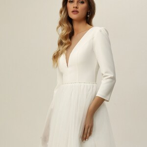 May include: A white wedding dress with a fitted bodice and a tulle skirt. The dress has a plunging neckline and three-quarter sleeves. The bodice has a beaded belt.