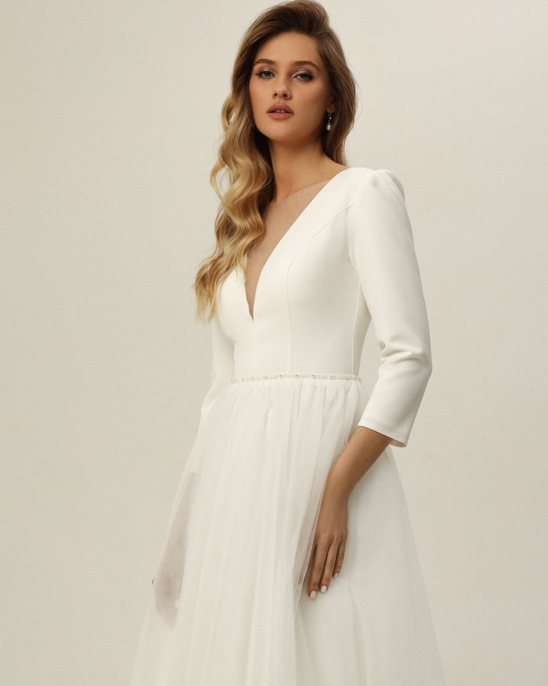 May include: A white wedding dress with a fitted bodice and a tulle skirt. The dress has a plunging neckline and three-quarter sleeves. The bodice has a beaded belt.