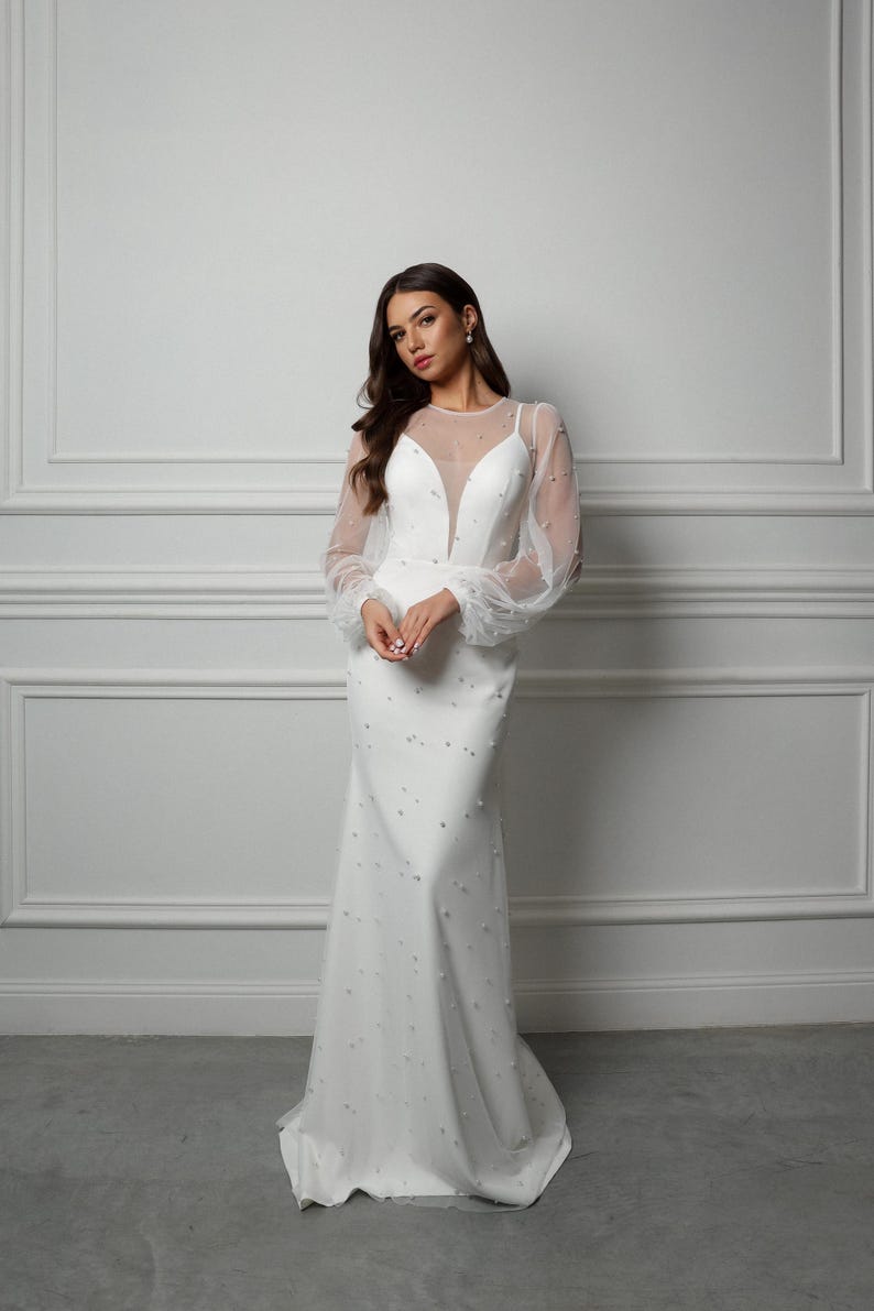 Op de afbeelding: Een witte bruidsjurk met een doorschijnende, met kralen bezette lange mouwen en een aansluitend lijfje. De jurk heeft een diepe V-hals en een zwierige rok.