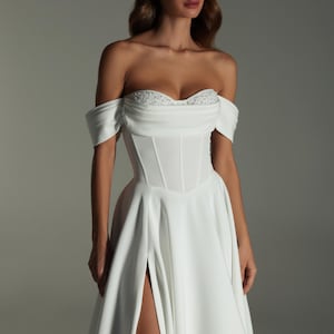 K&ouml;nnte beinhalten: Wei&szlig;es schulterfreies Brautkleid mit eng anliegendem Oberteil und flie&szlig;endem Rock. Das Oberteil hat einen Sweetheart-Ausschnitt mit zarten Verzierungen. Der Rock hat einen hohen Schlitz, der das Bein freilegt. Das Kleid ist aus einem glatten, eleganten Stoff.