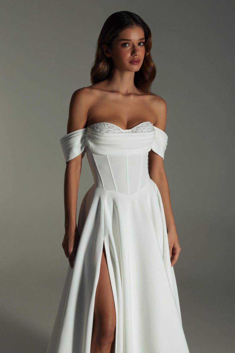 K&ouml;nnte beinhalten: Wei&szlig;es schulterfreies Brautkleid mit eng anliegendem Oberteil und flie&szlig;endem Rock. Das Oberteil hat einen Sweetheart-Ausschnitt mit zarten Verzierungen. Der Rock hat einen hohen Schlitz, der das Bein freilegt. Das Kleid ist aus einem glatten, eleganten Stoff.
