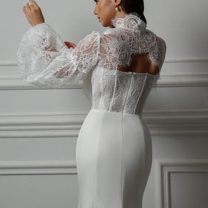 May include: A white lace wedding dress with a fitted bodice and a flowing skirt. The dress features a high neckline and long sleeves with a puff detail. The back of the dress has a keyhole opening.