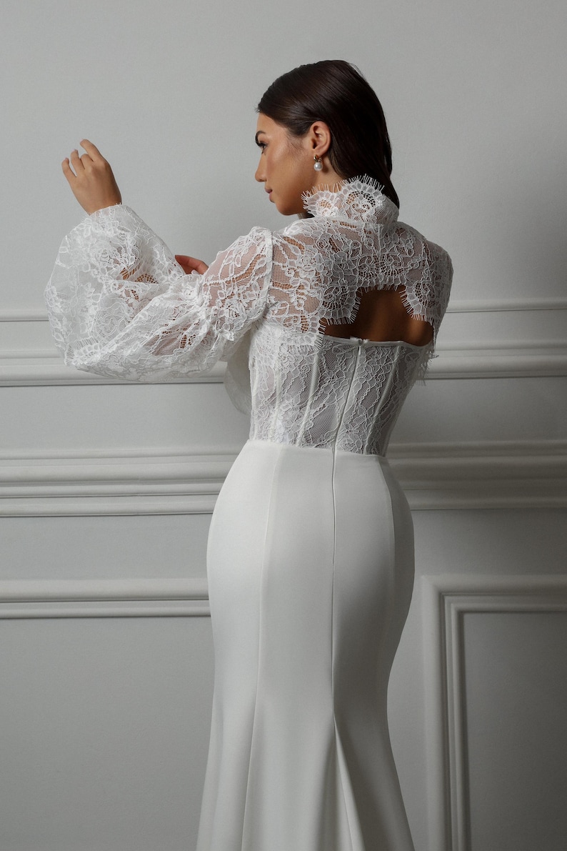 May include: A white lace wedding dress with a fitted bodice and a flowing skirt. The dress features a high neckline and long sleeves with a puff detail. The back of the dress has a keyhole opening.