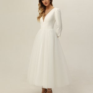 May include: A white wedding dress with a fitted bodice and a tulle skirt. The dress has a V-neckline and long sleeves. The bodice is adorned with a beaded belt.