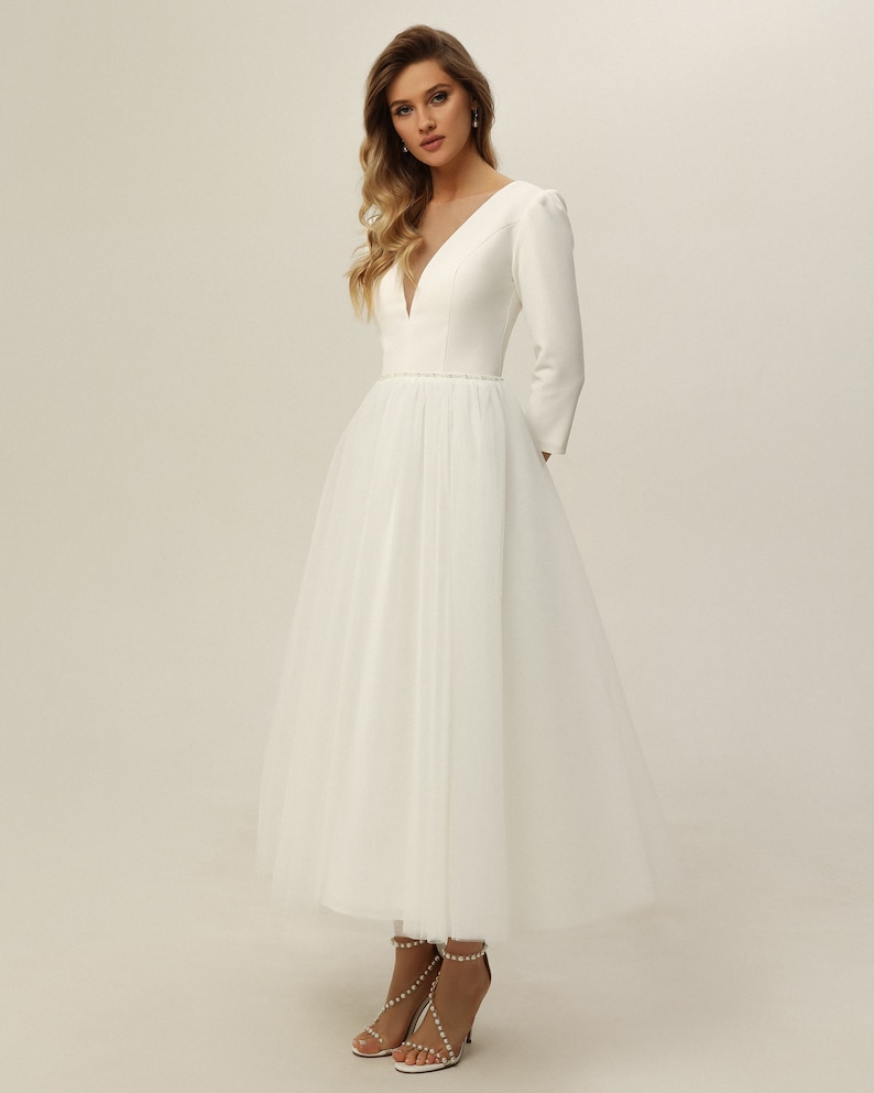 May include: A white wedding dress with a fitted bodice and a tulle skirt. The dress has a V-neckline and long sleeves. The bodice is adorned with a beaded belt.