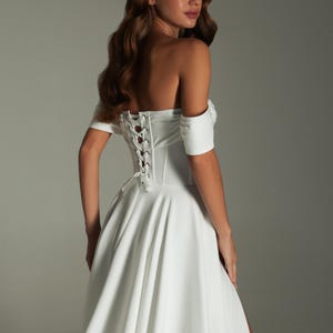 K&ouml;nnte beinhalten: Wei&szlig;es schulterfreies Brautkleid mit Korsettr&uuml;cken und flie&szlig;endem Rock. Das Kleid hat kurze &Auml;rmel und einen hohen Schlitz. Das Model blickt &uuml;ber die Schulter weg von der Kamera.