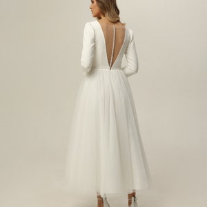 May include: A white wedding dress with a tulle skirt and long sleeves. The dress has a low back with a sheer panel and a row of pearl buttons.