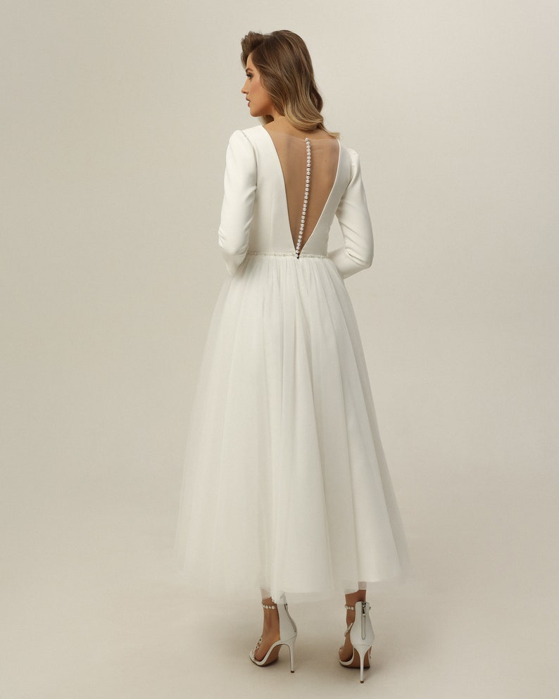 May include: A white wedding dress with a tulle skirt and long sleeves. The dress has a low back with a sheer panel and a row of pearl buttons.