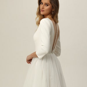 May include: A white wedding dress with a fitted bodice and a full tulle skirt. The dress has a low back with a pearl button closure.