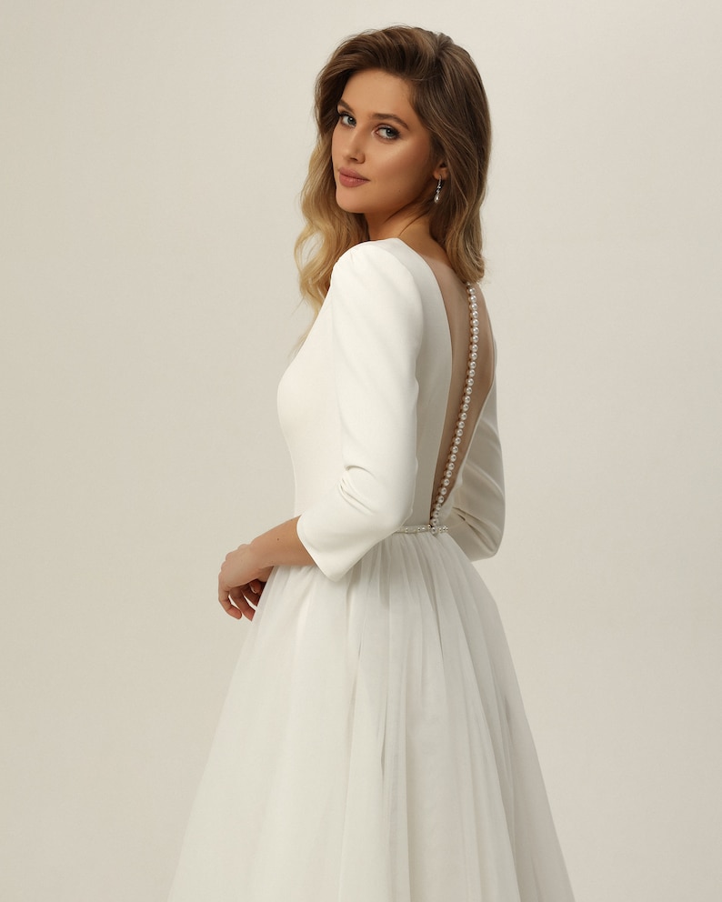 May include: A white wedding dress with a fitted bodice and a full tulle skirt. The dress has a low back with a pearl button closure.