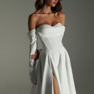 K&ouml;nnte beinhalten: Ein wei&szlig;es schulterfreies Kleid mit langen &Auml;rmeln und hohem Schlitz. Das Oberteil hat ein Korsett-Design und ist mit zarten Perlen verziert. Das Kleid ist aus flie&szlig;endem Stoff gefertigt.