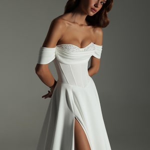 K&ouml;nnte beinhalten: Wei&szlig;es schulterfreies Hochzeitskleid mit eng anliegendem Oberteil und hohem Schlitz. Das Oberteil hat zarte Verzierungen. Das Kleid hat einen flie&szlig;enden Rock. Das Model steht vor einem grauen Hintergrund.