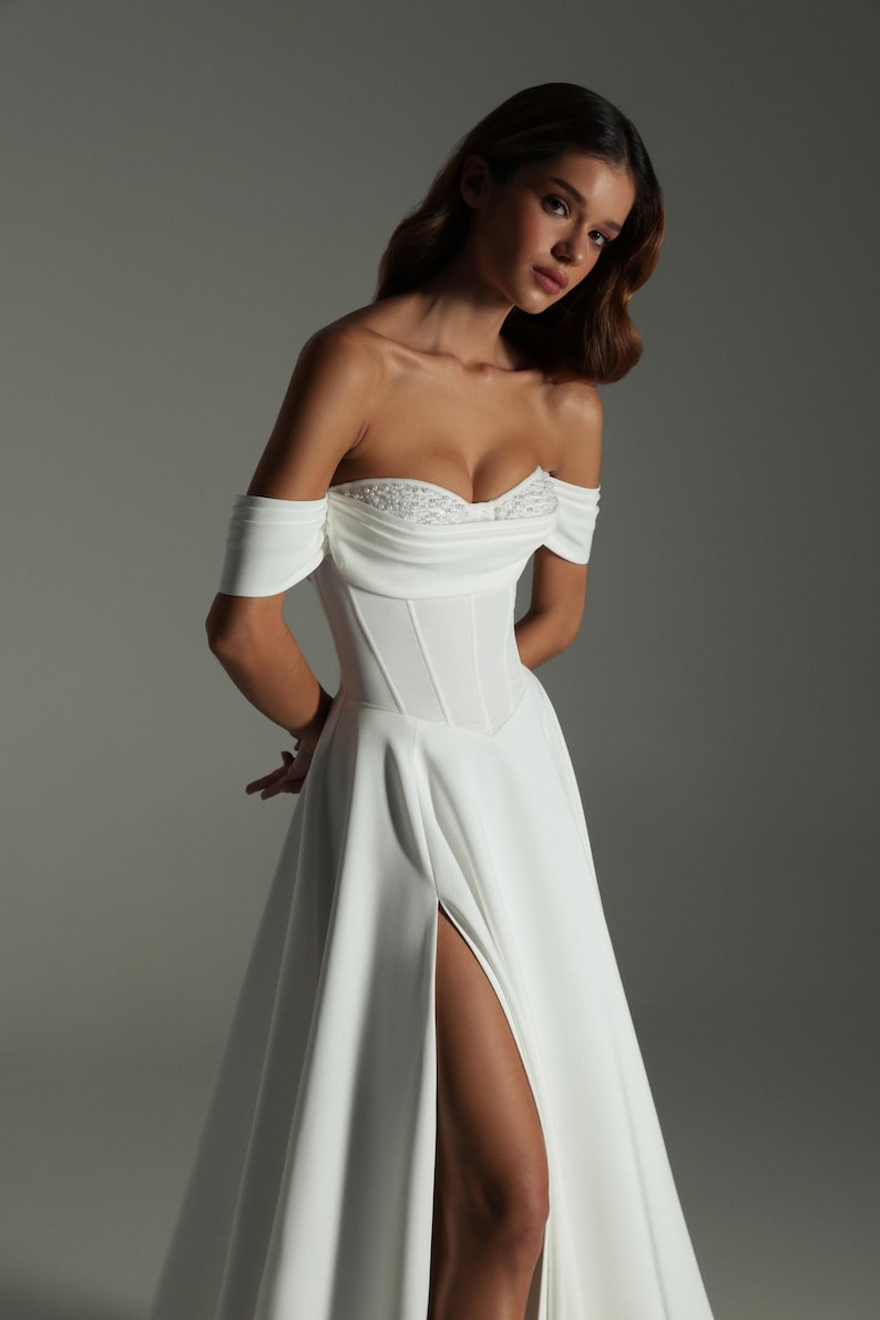 K&ouml;nnte beinhalten: Wei&szlig;es schulterfreies Hochzeitskleid mit eng anliegendem Oberteil und hohem Schlitz. Das Oberteil hat zarte Verzierungen. Das Kleid hat einen flie&szlig;enden Rock. Das Model steht vor einem grauen Hintergrund.