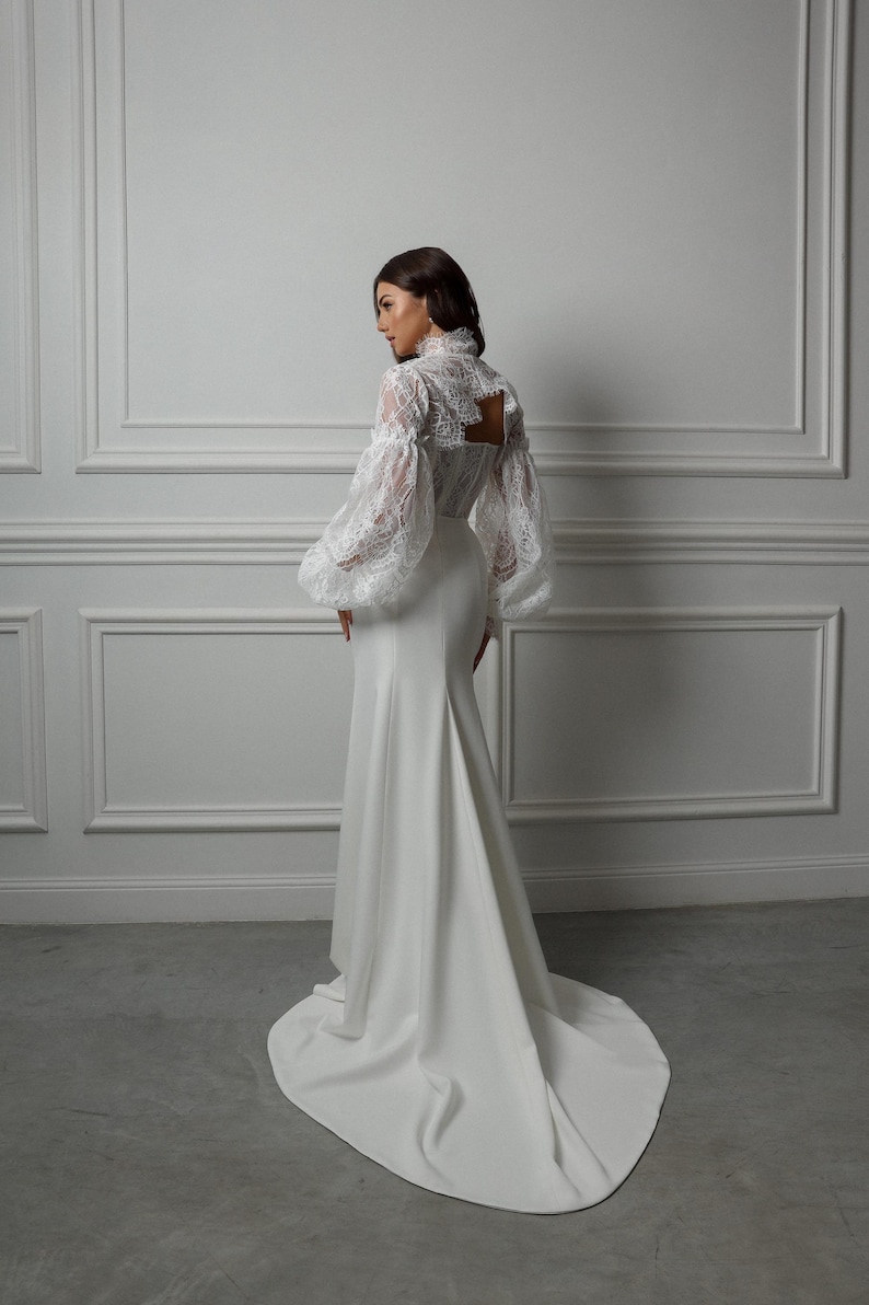 May include: A white wedding dress with a fitted bodice and a flowing skirt. The dress features a lace top with long, puffed sleeves.
