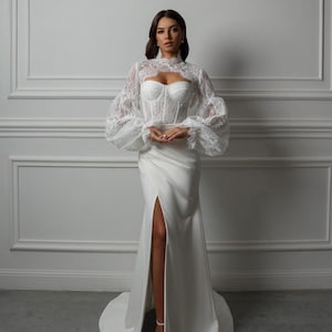 May include: A white lace top with long, puffed sleeves and a high neckline, worn over a white satin dress with a thigh-high slit.