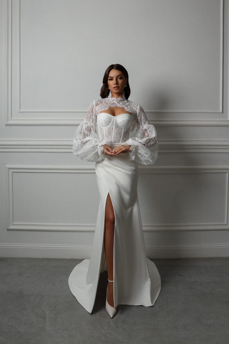 May include: A white lace top with long, puffed sleeves and a high neckline, worn over a white satin dress with a thigh-high slit.