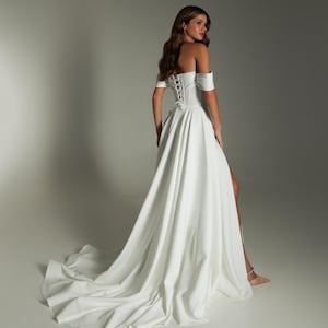 K&ouml;nnte beinhalten: Ein wei&szlig;es schulterfreies Hochzeitskleid mit Korsettr&uuml;cken und hohem Schlitz. Das Kleid hat eine flie&szlig;ende Schleppe und kurze &Auml;rmel. Das Model befindet sich in einem Studio.