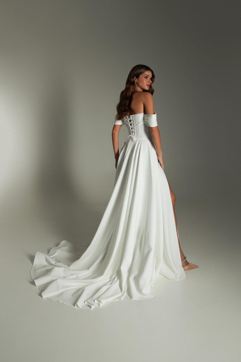 K&ouml;nnte beinhalten: Ein wei&szlig;es schulterfreies Hochzeitskleid mit Korsettr&uuml;cken und hohem Schlitz. Das Kleid hat eine flie&szlig;ende Schleppe und kurze &Auml;rmel. Das Model befindet sich in einem Studio.