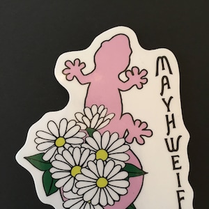 Op de afbeelding: Een sticker met een roze hagedisachtig figuur met een boeket madeliefjes. De madeliefjes hebben witte bloemblaadjes en gele centra. De tekst "MAY WEIRD" staat verticaal aan de rechterkant.