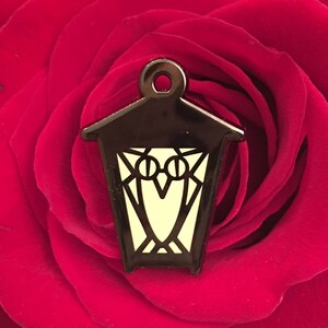 May include: A black metal lantern charm with a glow-in-the-dark owl design. The lantern has a pointed roof and a small loop at the top for hanging.