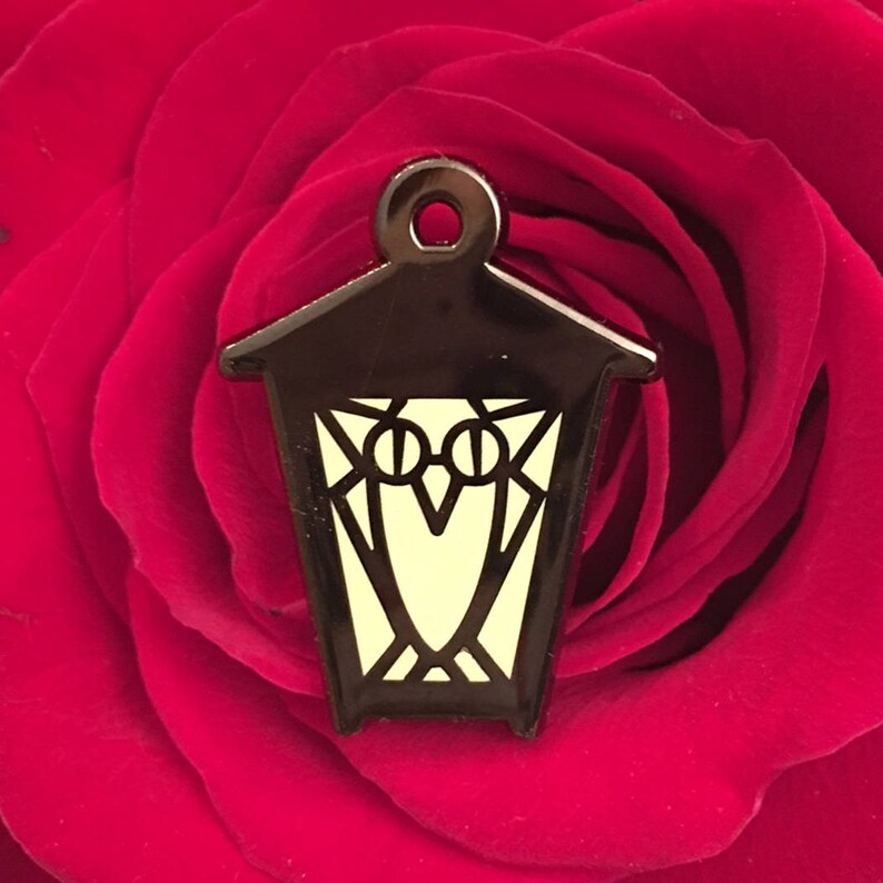 May include: A black metal lantern charm with a glow-in-the-dark owl design. The lantern has a pointed roof and a small loop at the top for hanging.