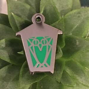 May include: A silver metal lantern pin with a green enamel design. The lantern has a pointed roof and a rectangular body.