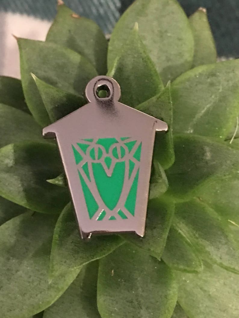 May include: A silver metal lantern pin with a green enamel design. The lantern has a pointed roof and a rectangular body.