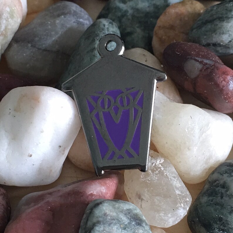 May include: A silver metal lantern pin with a purple enamel design. The lantern has a geometric pattern and a pointed top.