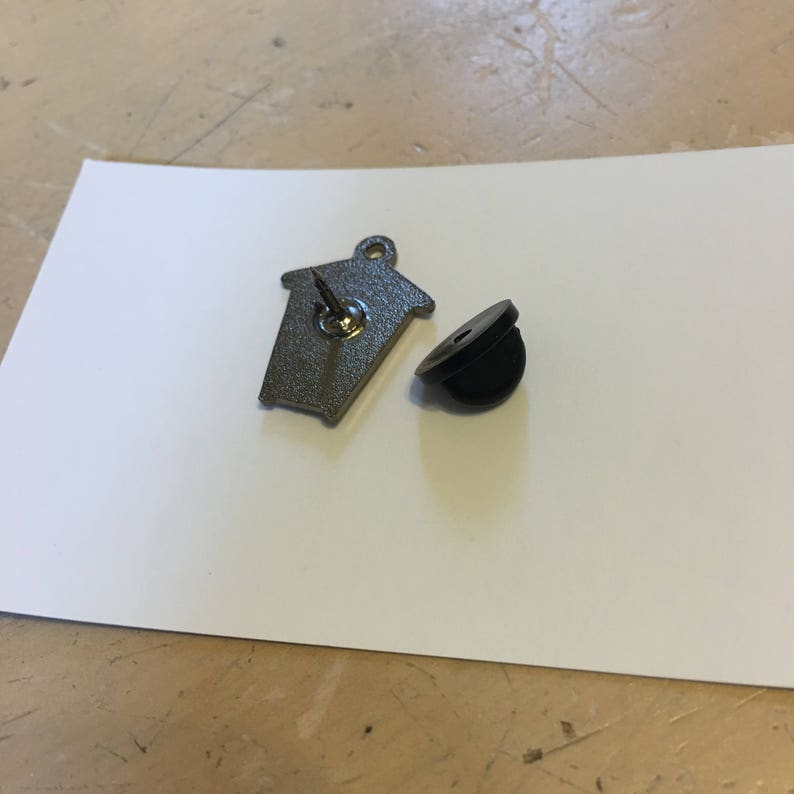 May include: A silver metal pin in the shape of a house with a black rubber backing. The pin is on a white surface.