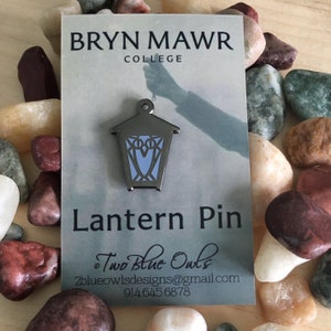 May include: A silver lantern pin with a blue owl design. The pin is on a white card with the text "Bryn Mawr College" and "Lantern Pin". The card also includes the text "Two Blue Owls" and contact information.