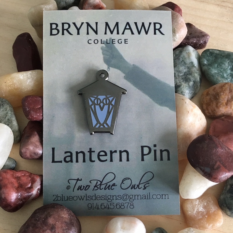 May include: A silver lantern pin with a blue owl design. The pin is on a white card with the text "Bryn Mawr College" and "Lantern Pin". The card also includes the text "Two Blue Owls" and contact information.