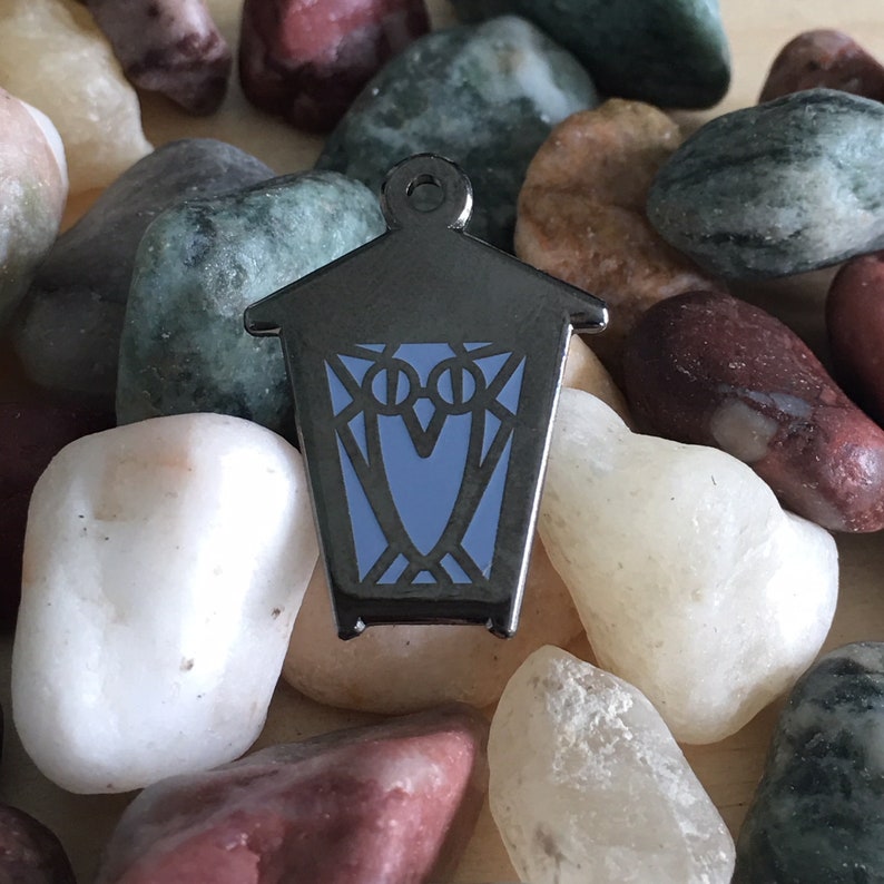 May include: A silver metal lantern pin with a blue owl design. The lantern has a pointed roof and a rectangular body with a window on each side. The owl is facing forward with its wings spread out.